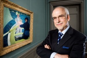 An older man in a suit and striped tie stands with arms crossed, smiling, in front of a framed painting of people by the water, in a room with teal walls.