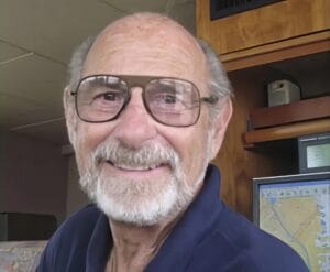 An older man with glasses and a white beard, smiling, stands indoors by wooden shelves and a computer monitor displaying a map.