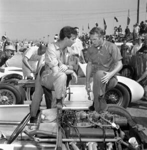 Two men in racing suits talk beside a vintage race car, one kneeling on its tire; people, cars, and spectators fill the racetrack.