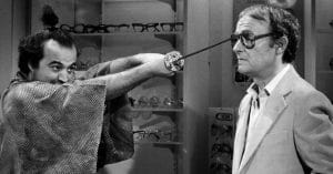 A man in a kimono holds a sword, with its tip pressed against the glasses of another man in a suit and glasses, who appears calm; eyeglasses are displayed on shelves behind them.