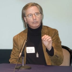 A person with light brown hair and glasses speaks at a table with a microphone, wearing a brown jacket and name tag: Burley Easley.