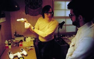 Two men talk in a dim room. One in glasses and a yellow shirt holds a model spaceship; the other listens by a cluttered worktable.