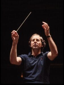A man wearing glasses and a dark shirt is conducting, holding a baton in one hand and raising the other hand, against a dark background.