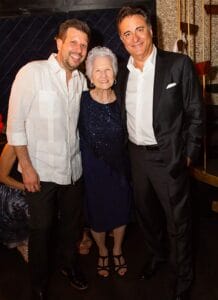 Three people smile, standing indoors; an elderly woman in navy stands between two men. Background has dark walls and gold accents.