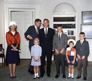 A vintage photo of President Kennedy, Scott and Rene Carpenter, and four four children in the White House with bookshelves behind them.
