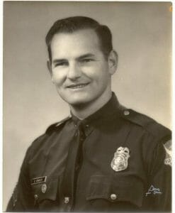 A smiling male police officer in uniform poses for a black and white portrait against a plain, light background. Badge and patches visible.