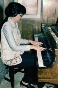 A woman with short dark hair and glasses plays a Steinway grand piano, seated on a chair in a wood-paneled room, wearing casual attire.