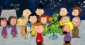 A group of Peanuts cartoon characters, including Charlie Brown and Snoopy, stand around a decorated Christmas tree outdoors at night, smiling and celebrating under a starry sky.