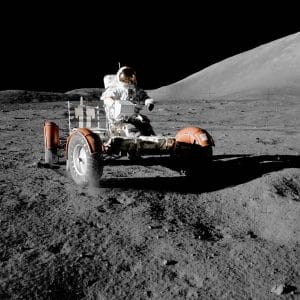 An astronaut wearing a spacesuit drives a lunar rover across the rocky, gray surface of the Moon under a black sky, with a hilly lunar landscape in the background.