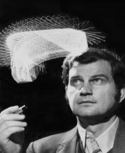 A man in a suit holds a pen, looking upward thoughtfully. Above him swirls a geometric light pattern on a dark background, evoking creativity.