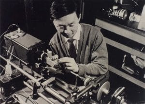 A man in a cardigan and tie adjusts a piece of scientific equipment on a laboratory workbench, surrounded by various instruments and tools.