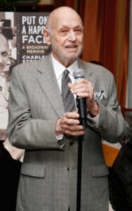 An older man in a gray checked suit holds a microphone and speaks, with a poster for “Put On a Happy Face: A Broadway Memoir” behind him.
