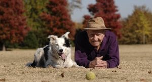 An elderly man wearing a brown hat and purple shirt lies on grass next to a black and white dog. Both are looking forward, with trees showing autumn colors in the background and a tennis ball in the foreground.