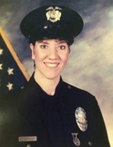 A smiling woman in a police uniform and hat poses for a formal portrait in front of a U.S. flag. She has short brown hair and wears a badge and nameplate on her uniform.