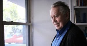 An older man with gray hair, wearing a blue shirt and dark sweater, stands by a window indoors, smiling as he looks outside.