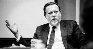 A bearded man in a suit and tie gestures with his left hand while speaking, seated at an indoor table. Black and white photo.