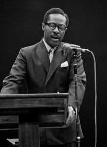 A period black & white photo from the 1960s shows a man wearing glasses and a suit as he speaks into a microphone while standing at a lectern against a dark background.
