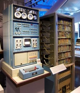 A vintage computer system with multiple panels of switches, dials, lights, and a small display screen, alongside an open cabinet revealing complex wiring and circuits. The setup is displayed in a museum setting.