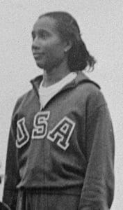 Black-and-white photo of a female athlete wearing a USA track suit, standing and looking to the side. Her hair is pulled back, and she appears to be outdoors.