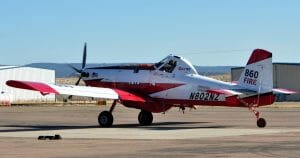 A red and white single-engine firefighting plane, 860 FIRE, is on the tarmac by hangars. Registration N802NZ is visible.