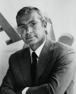 Black and white portrait of an older man with glasses and graying hair, in a suit and tie, looking neutrally at the camera. Abstract shapes behind.