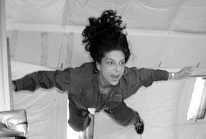 A woman with long dark hair floats in mid-air inside a space simulation, wearing a jumpsuit and looking amazed, experiencing weightlessness.