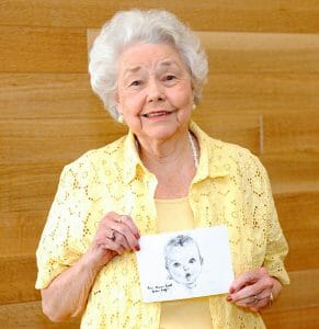 Elderly woman with white hair in a yellow blouse smiles, holding a baby pencil sketch, standing before a wooden wall background.