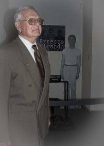 An older man in a suit stands in front, with a black-and-white cutout of a casually dressed young man and an Ernesto Miranda sign behind him.