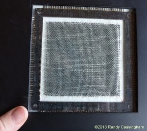 A persons hand holds a square glass frame containing a piece of fine mesh fabric, with measurement markings along the edges. The photo is credited to Randy Cassingham, ©2018.