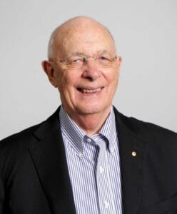 An older man with glasses, a bald head, and a friendly smile wears a black blazer over a striped shirt against a plain light background.