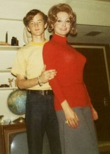 A teenage boy in a yellow shirt stands with his hand on the waist of a smiling woman in a red turtleneck and gray skirt. They pose indoors near a wooden shelf, a globe, and a TV.