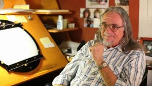 Smiling older man with long gray hair and glasses sits by a drawing desk cluttered with papers, photos, and art supplies.