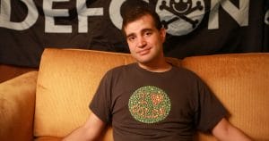 A person with short dark hair smiles slightly while sitting on a tan couch in front of a DEFCON banner, wearing a dot-patterned shirt.