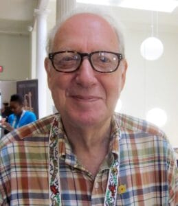 An older man with glasses and a plaid shirt smiles at the camera indoors, with round lights and columns visible behind him.