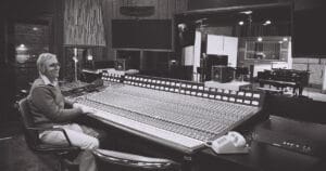 A man sits at a large audio mixing console in a studio control room, equipment and monitors around, viewing a grand piano and drums.