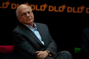 An older man in a dark suit and light blue shirt sits on stage with headset mic, 'DLD' logos behind him. He looks thoughtful.