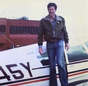 A man in a flight jacket and blue jeans stands on the wing of a small airplane, parked on the ground. There is a building in the background.