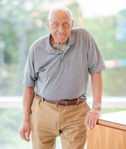 An elderly man with short white hair, in a gray polo and khaki pants, leans on a wooden lectern indoors, smiling at the camera.