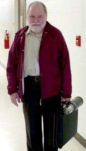 An older man with a white beard, red jacket, and dark pants holds a briefcase and thermos in a school hallway with white walls.