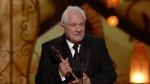 An older man in a black tuxedo stands at a microphone, holding an Oscar statuette, with a gold and brown backdrop behind him.