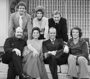 The cast and creators of the Newhart show, five men and two women, pose in two rows indoors; four on a couch, three standing, all smiling at the camera.