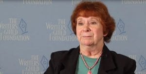 A woman with short reddish hair wearing a dark blazer, green top, and pink necklace sits in front of a blue backdrop with The Writers Guild Foundation logo and text.