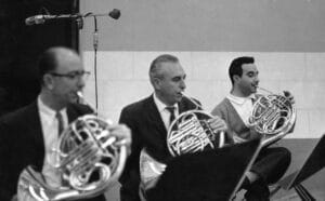 Three men play French horns in a row, focused, with music stands and a microphone visible; the black and white scene suggests a recording session.