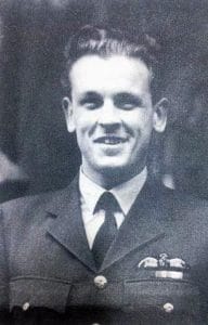 Black and white photo of a young man in a military uniform, smiling at the camera. He has short, neatly styled hair and is wearing a jacket with buttons and a winged badge on the chest.
