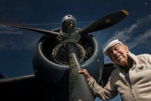 An elderly man wearing glasses, a white cap, and a light jacket stands next to a large vintage airplane, which he holds by the propeller, looking up and smiling against a blue sky.