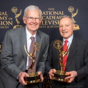 Two older men in suits smile and hold Emmy awards in front of a backdrop reading The National Academy of Television Arts & Sciences.