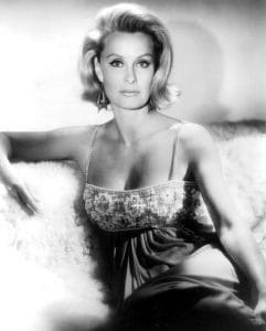 Black and white photo of a woman with shoulder-length blonde hair, wearing a sleeveless, embellished dress, sitting on a soft, furry surface and looking directly at the camera with a neutral expression.