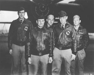Five men in military uniforms and leather flight jackets stand together in front of a bomber, posing for a group photo. Four wear caps, and all display serious expressions. A wing and engine are visible behind them.