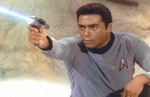 A man in a blue Star Trek uniform kneels and aims a phaser emitting a blue beam, with a focused expression, against a rocky background.
