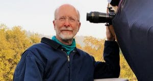 An older man with glasses and a white beard in a blue jacket stands outdoors by a large telescope, trees and sky behind him.